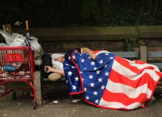 NEW YORK, NY - SEPTEMBER 10: A homeless man sleeps under an American Flag blanket on a park bench on September 10, 2013 in the Brooklyn borough of New York City. As of June 2013, there were an all-time record of 50,900 homeless people, including 12,100 homeless families with 21,300 homeless children homeless in New York City. (Photo by Spencer Platt/Getty Images)